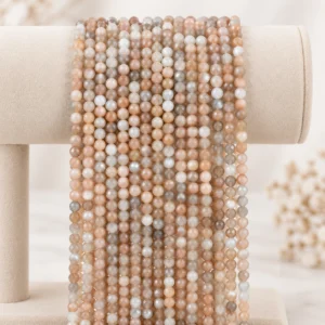 Soft beige, peach, white, and grey faceted gemstone bead strands draped over a cream velvet display stand.